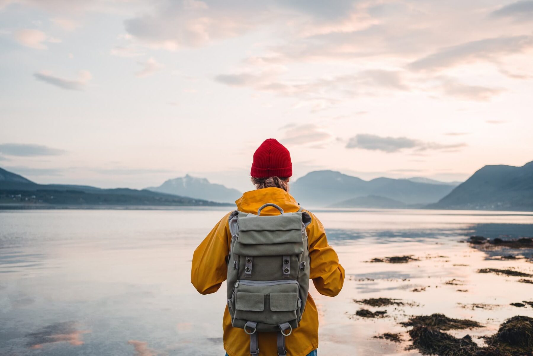 A person wearing a yellow jacket, red beanie, and backpack stands by a calm lake, looking toward distant mountains under a cloudy sky at sunset.