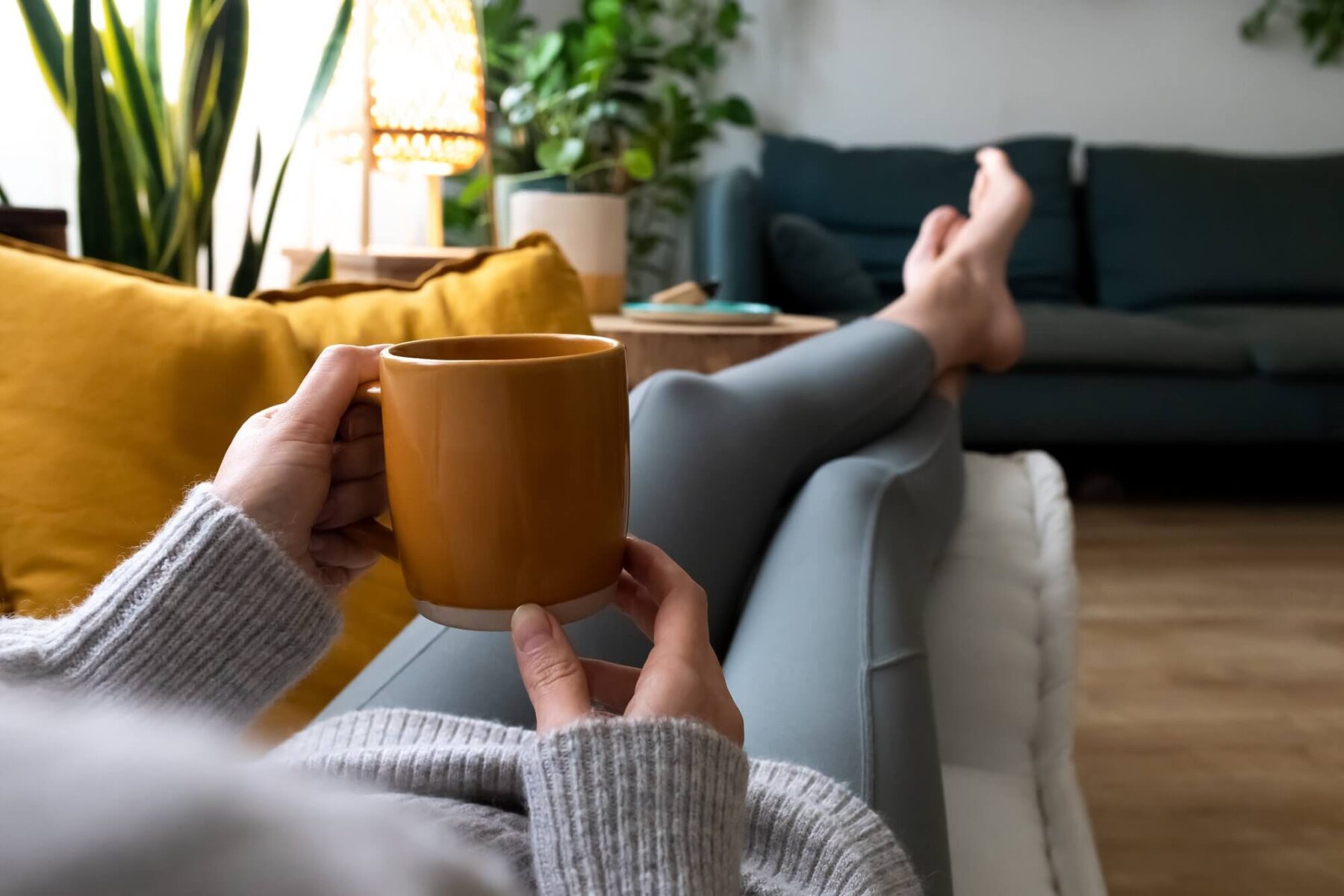 A person in a cozy sweater relaxes on a sofa, holding a yellow mug with legs stretched out. The room features green plants, a yellow cushion, and a soft, inviting atmosphere.