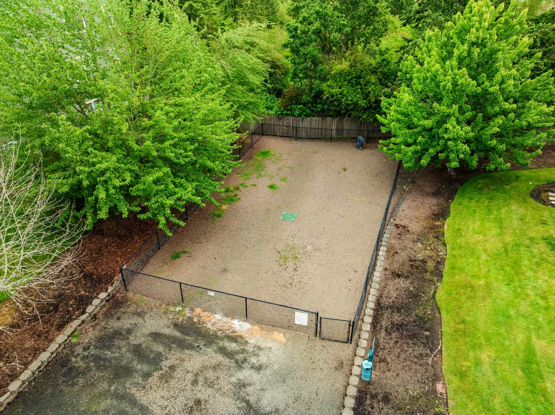 Aerial view of a fenced, rectangular dirt dog park area surrounded by lush green trees and grass.