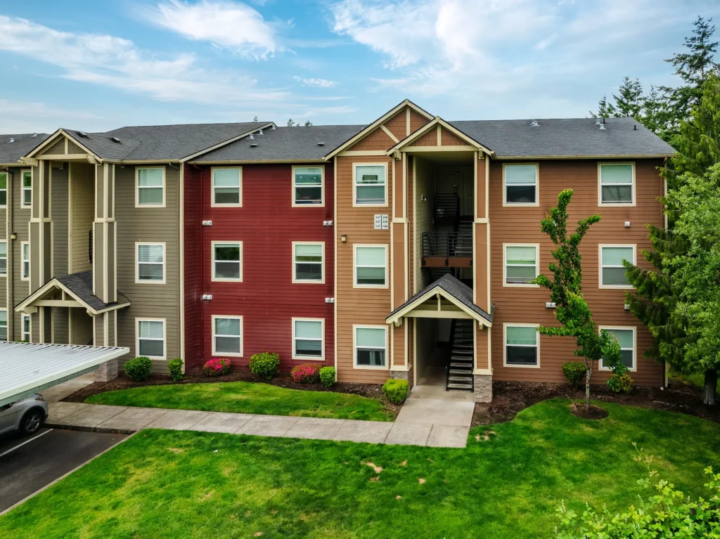 Three-story apartment building with red and beige siding, surrounded by green lawn and trees. A path leads to a central staircase.