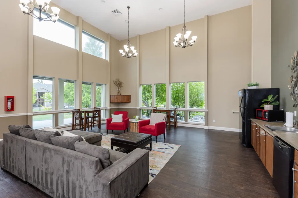 Spacious clubhouse with high ceilings, large windows, and chandeliers. Features a gray couch, red chairs, and a kitchenette with modern appliances.