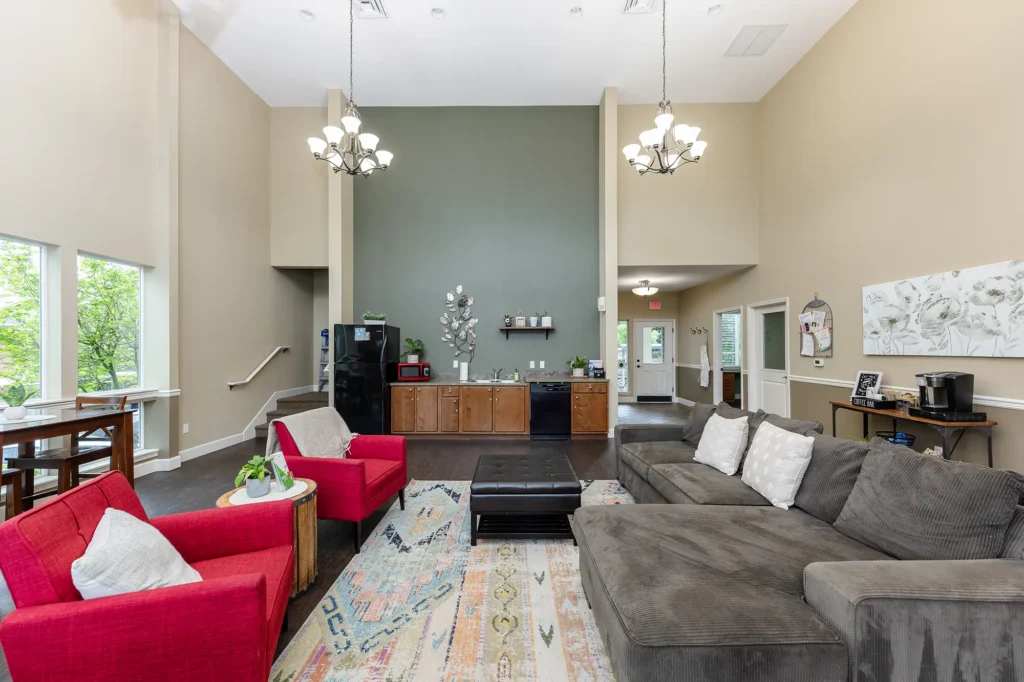 Spacious clubhouse with high ceilings, large windows, and chandeliers. Features a gray couch, red chairs, and a kitchenette with modern appliances.