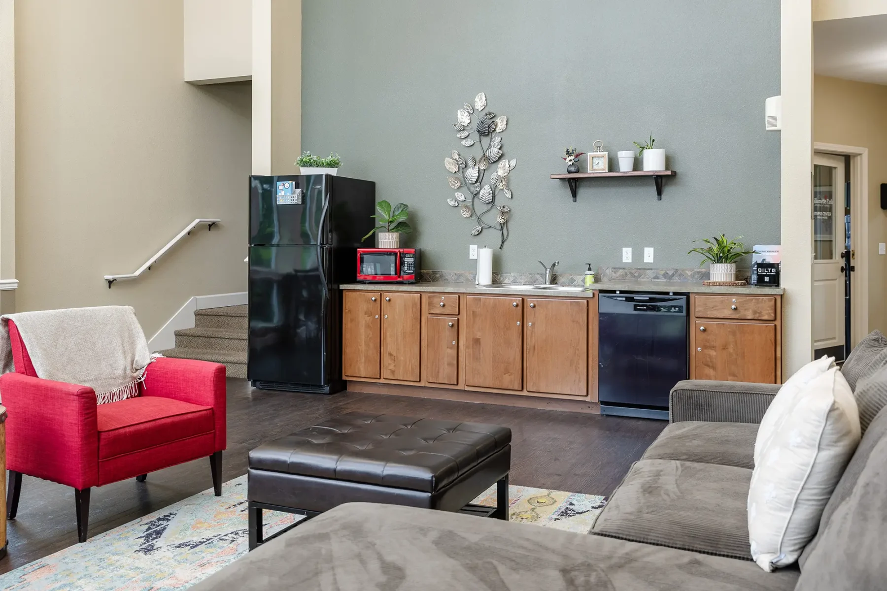 Clubhouse with a red armchair, gray sectional sofa, and black ottoman. Kitchenette space has wood cabinets, black appliances, and wall art.