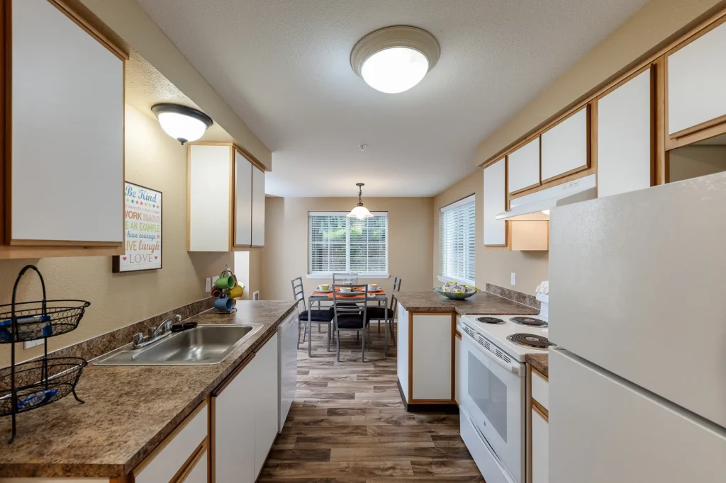 Kitchen with white cabinets, brown countertops, and wood-textured flooring. Dining area with table set for four near two large windows.