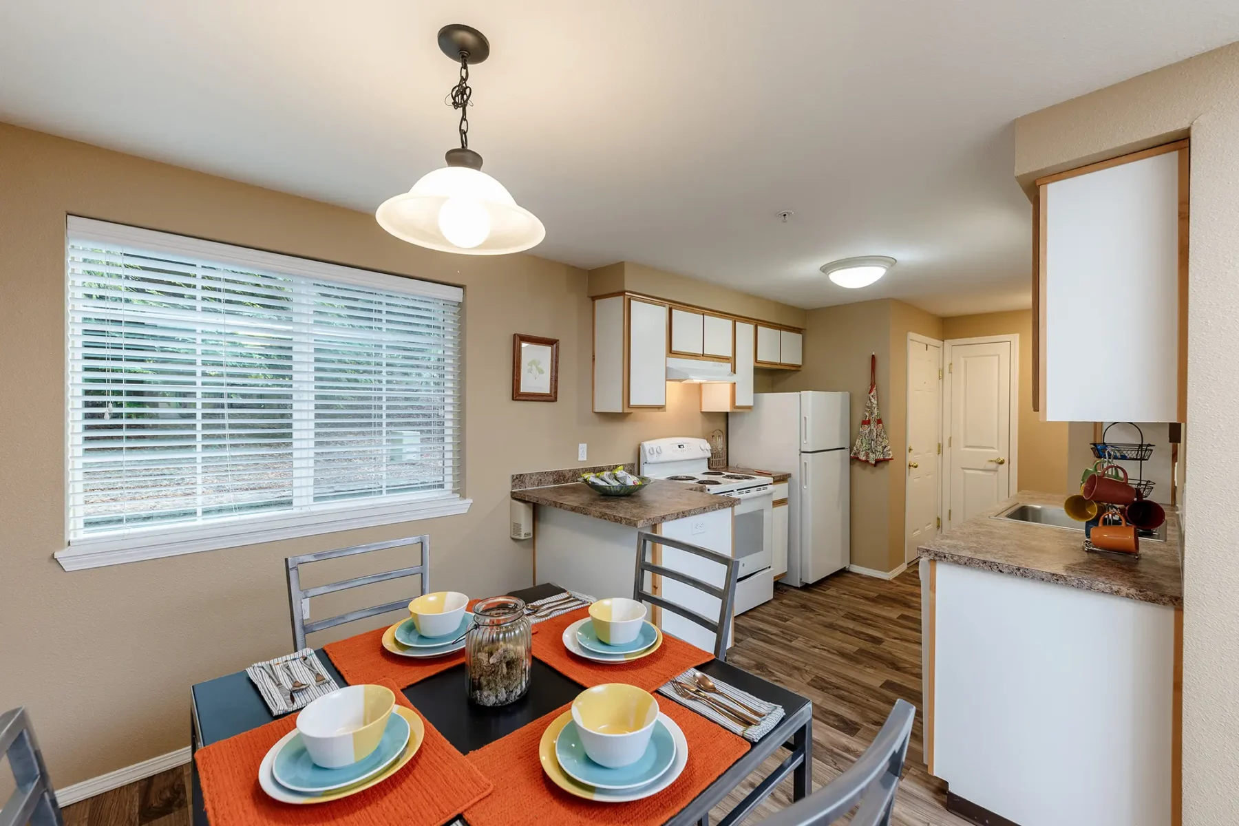 Kitchen with white cabinets, brown countertops, and wood-textured flooring. Dining area with table set for four near two large windows.