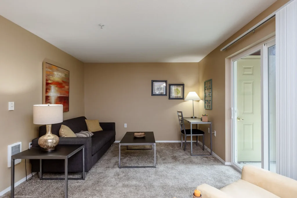 A cozy living room with beige walls, a dark sofa, and abstract artwork. Small desk with a lamp, and a glass door leading outside.