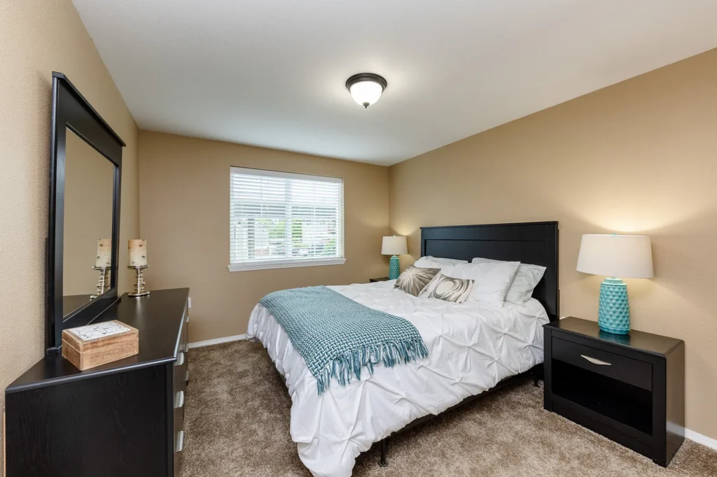 A bedroom with beige walls, large bed with white bedding and teal accents, plus a dresser with a mirror and nightstands with lamps.