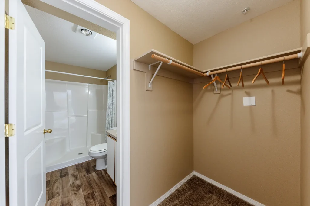 Spacious closet with wooden rods and hangars next to bathroom with standing shower, toilet, and sink.
