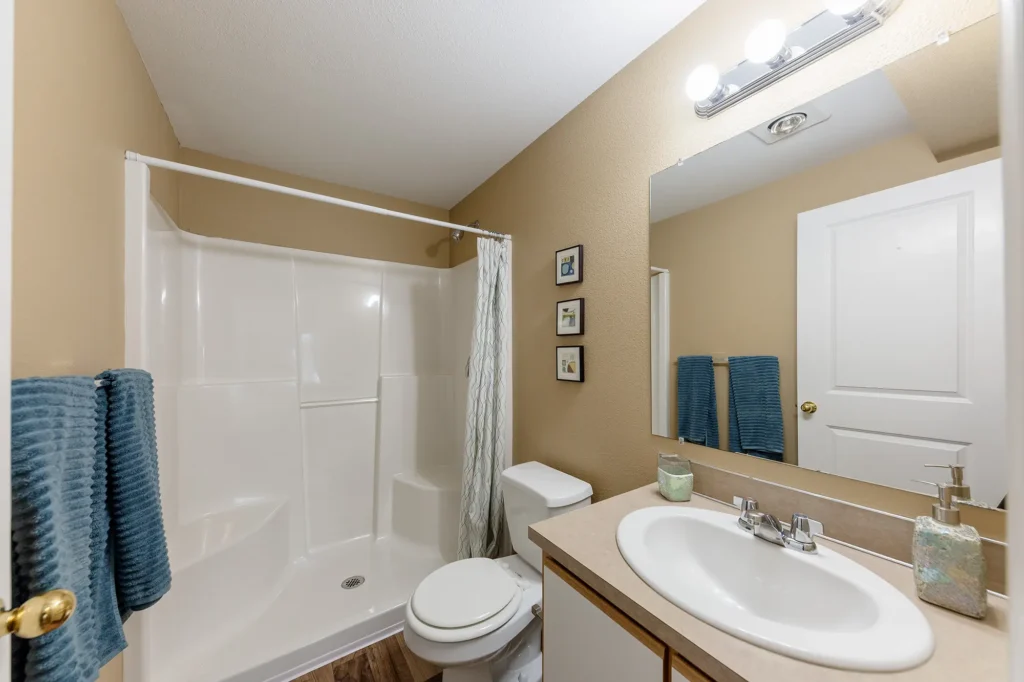 Bathroom with beige walls, a white standing shower and curtain, toilet, sink with toiletries, and mirror above.