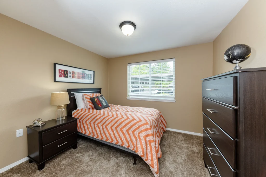 Bedroom with carpet and beige walls, window with blinds, and single bed with dresser and nightstand.