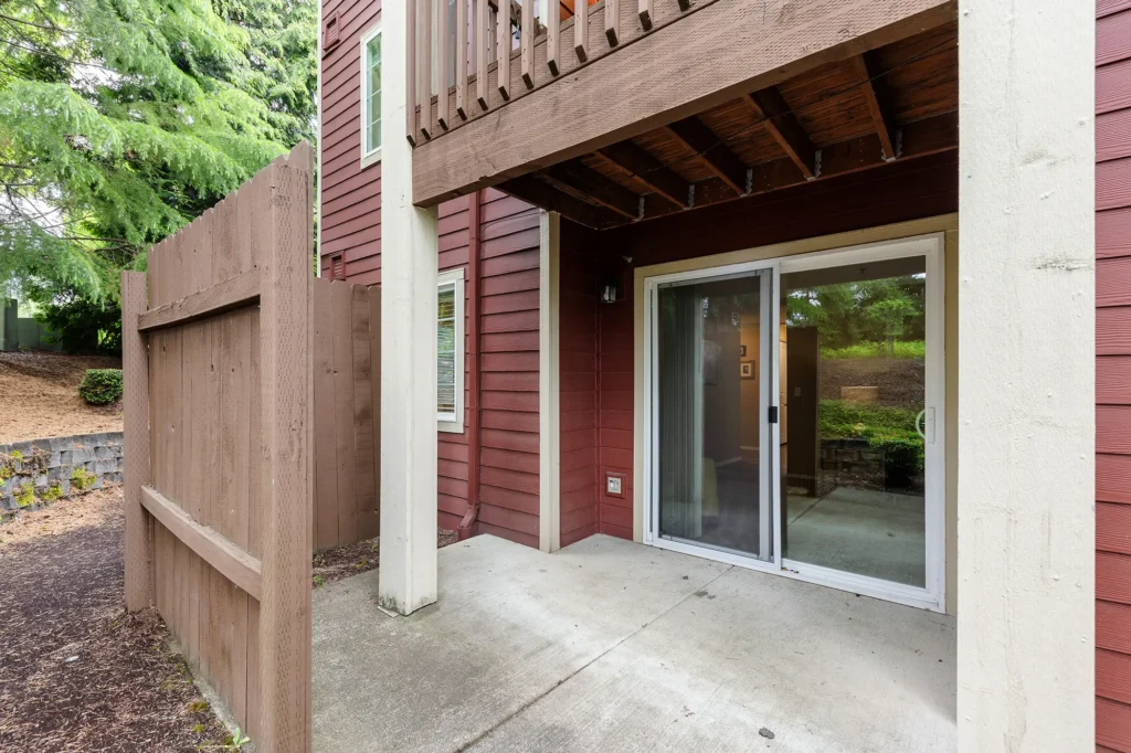 Outdoor patio space with a sliding glass door, brown wooden fence, and overhead balcony.