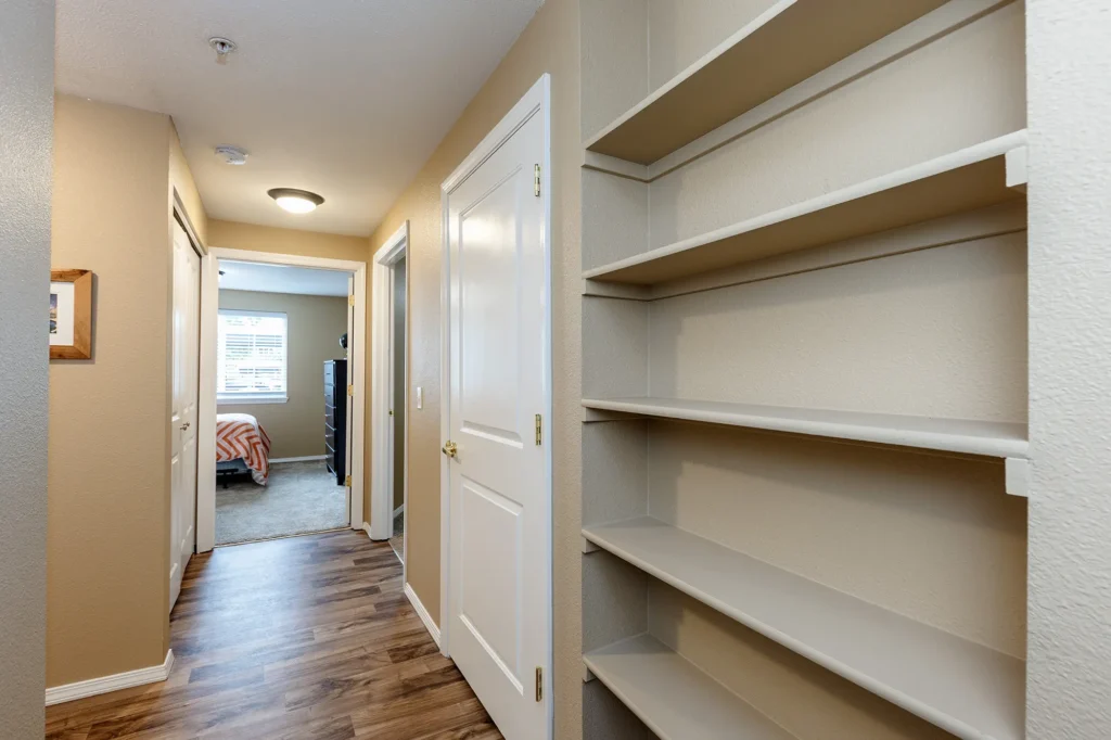 Hallway with wooden floor, beige walls, and white trim features built-in shelves on the right and a view into a carpeted bedroom.