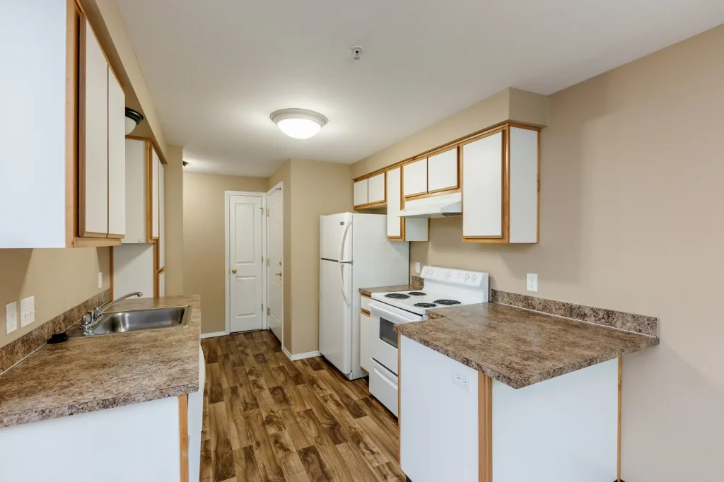 Kitchen with wooden floors, tan walls, and granite-style countertops. Includes white appliances, cabinets, and overhead lighting.