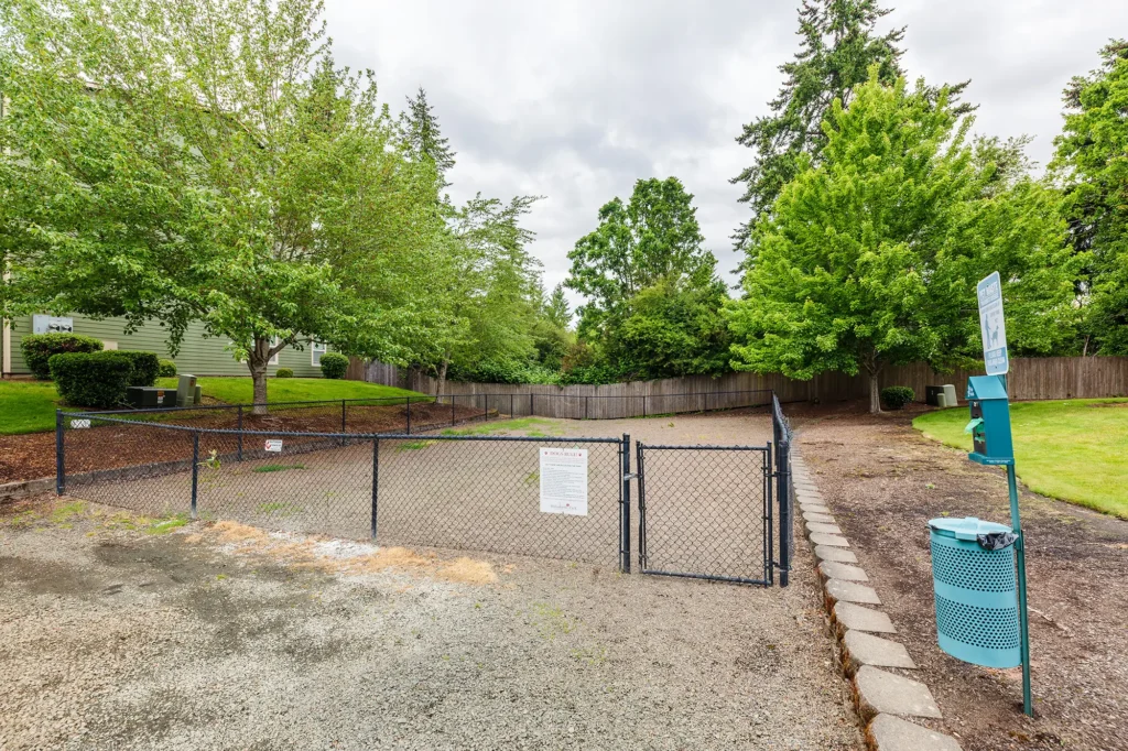 Fenced in dog park with trash can, surrounded by lush greenery.