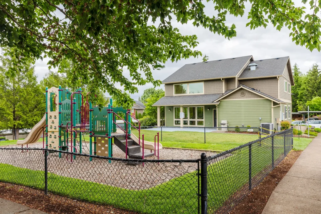A playground with colorful slides, climbing structures, and a safety fence sits beside a green, two-story leasing center. A tree and path are in the foreground.