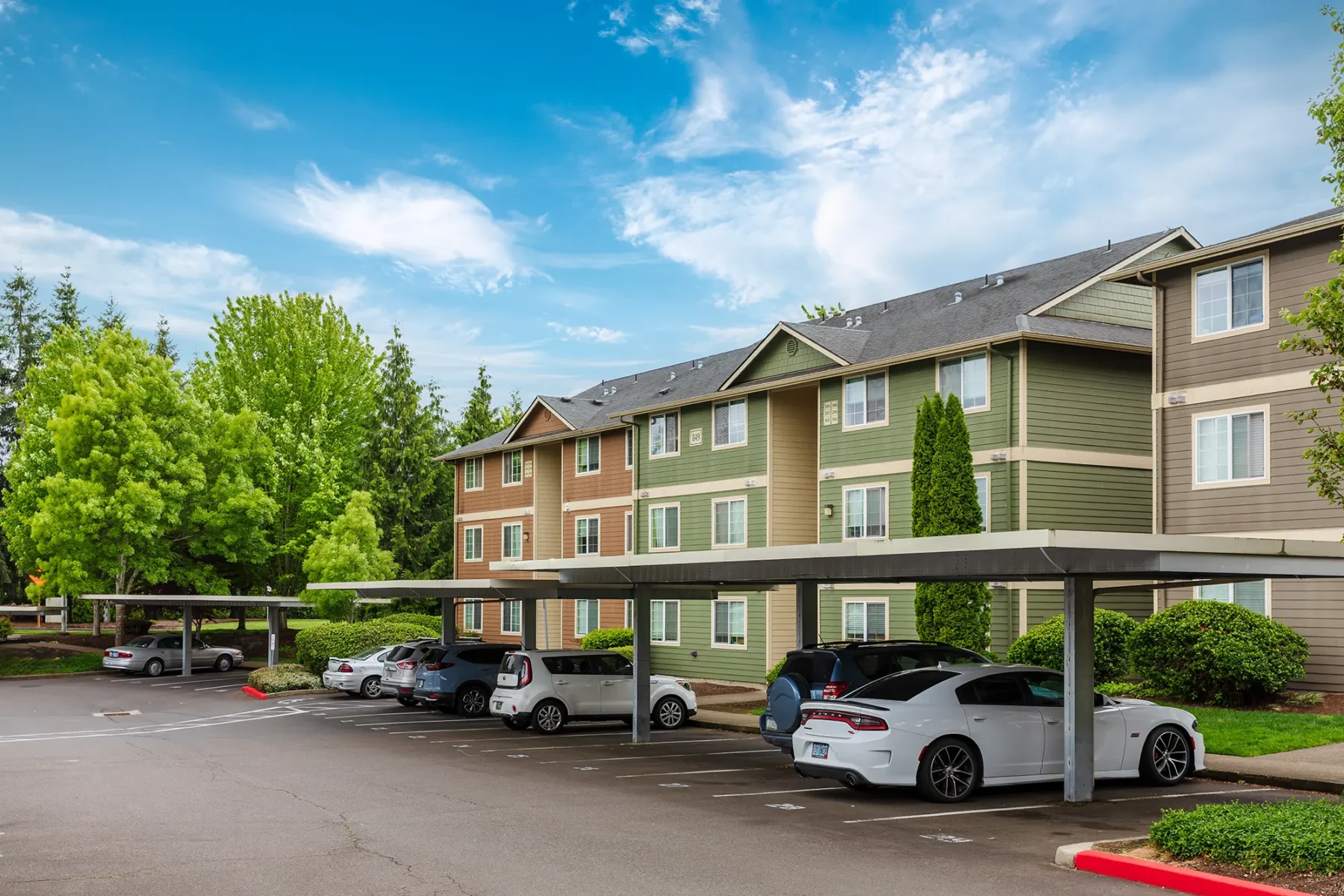 Modern apartment complex with beige, brown, and green siding. Covered parking with several cars beneath.