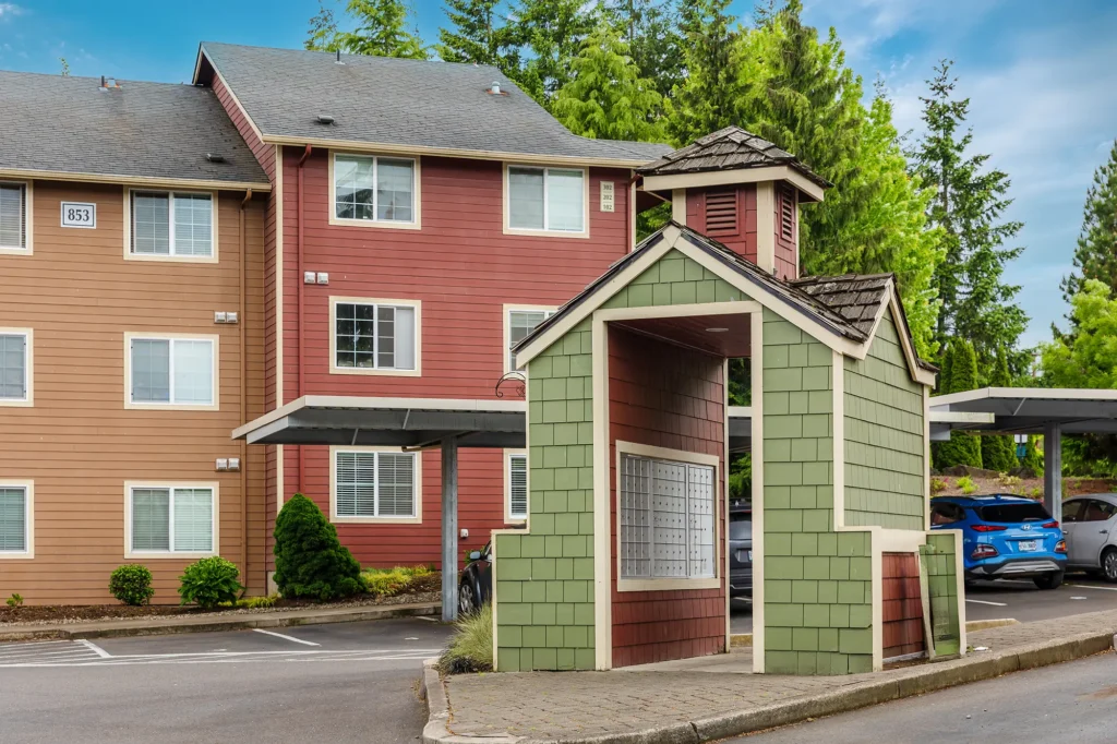 Suburban apartment complex with cars parked under a metal carport. Red and brown buildings with a small covered mailbox space.