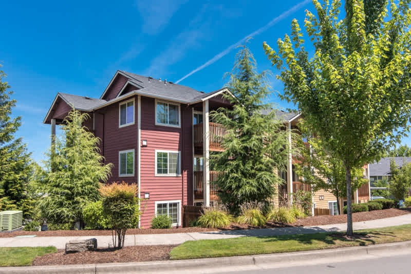 Three-story red, pet friendly apartment building with balconies, surrounded by green trees and shrubs on a sunny day. A sidewalk and street run in front of the building under a clear blue sky.