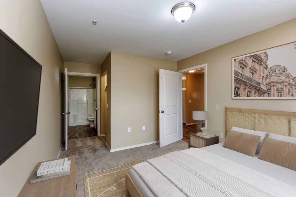 A modern bedroom with beige walls, a bed with neutral bedding, a large wall-mounted TV, a nightstand, and a framed photo of a building. The room has an open door leading to a bathroom and a hallway.