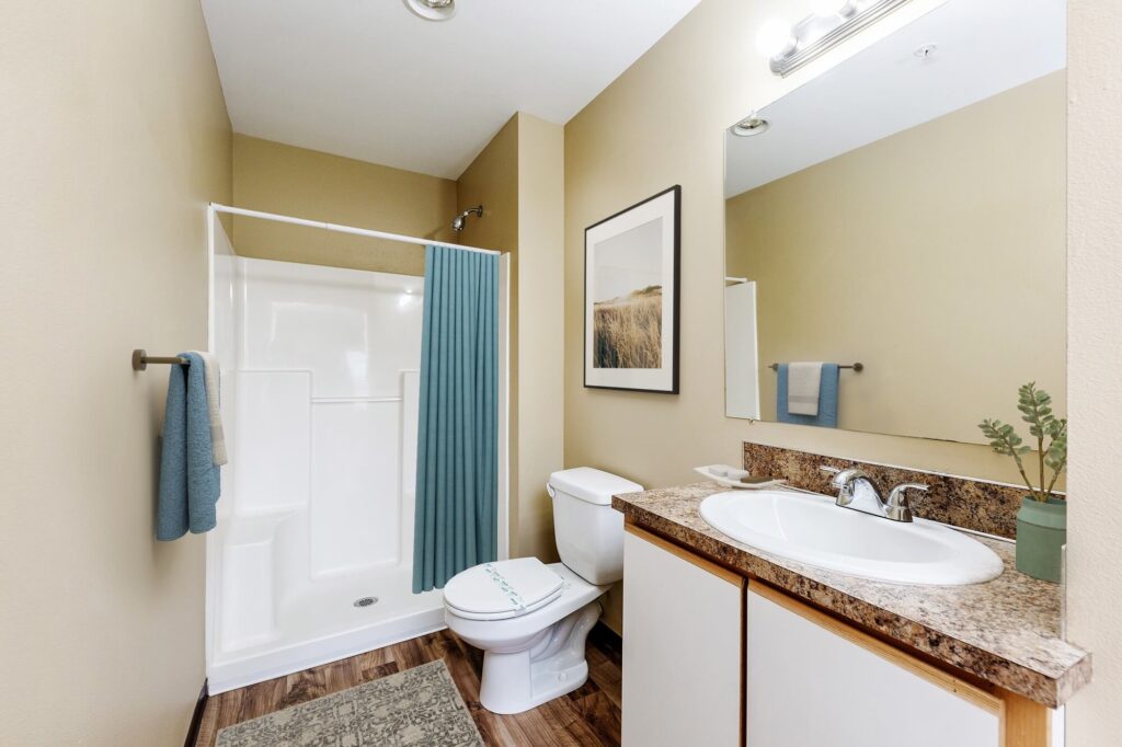 A clean bathroom with beige walls, a shower with a blue curtain, a toilet, a sink with a marble countertop, a wall mirror, towels, and a framed nature photo above the toilet.