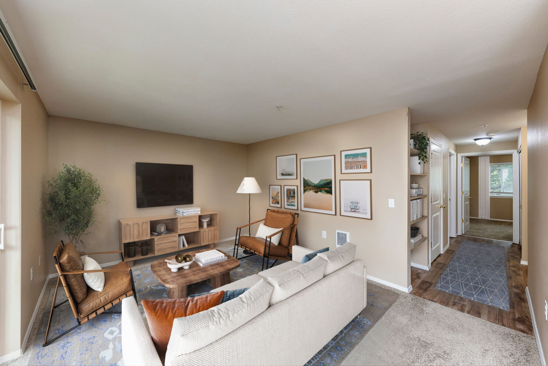 A modern living room with beige walls, a light sofa, two brown chairs, a wooden coffee table, TV on the wall, framed art, a floor lamp, a potted plant, and a hallway leading to other rooms with wood and carpet flooring.