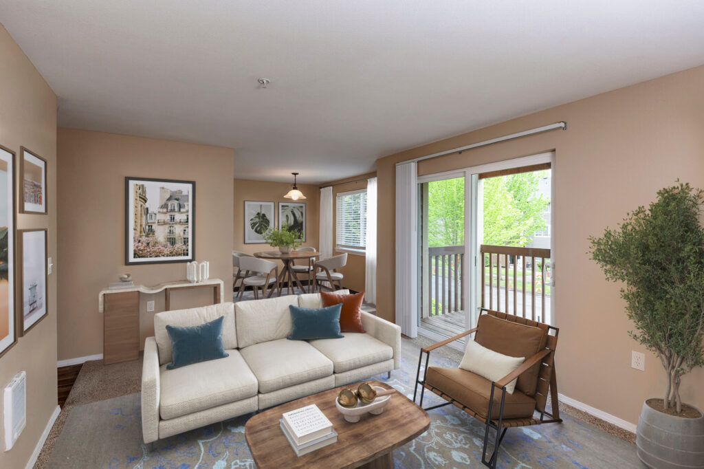 A modern living room with tan walls, a white sofa with blue pillows, a wooden coffee table, and a leather chair. Large windows let in natural light and offer a view of greenery outside. A dining area is visible in the background.