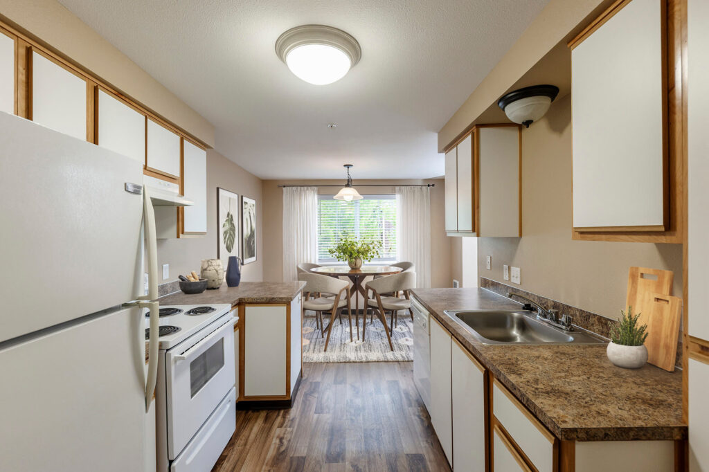 A bright galley kitchen with white appliances and brown countertops leads to a dining area with a round table, four chairs, and a large window with sheer curtains letting in natural light.