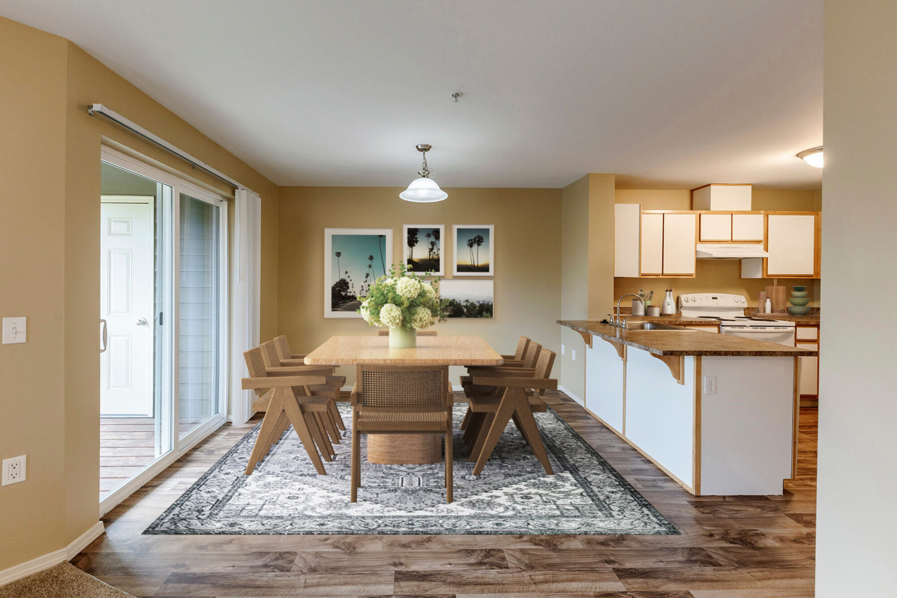 A dining area with a wooden table, six chairs, a vase of flowers, a patterned rug, wall art, and an open kitchen with white cabinets and appliances. Sliding glass doors lead to a patio.