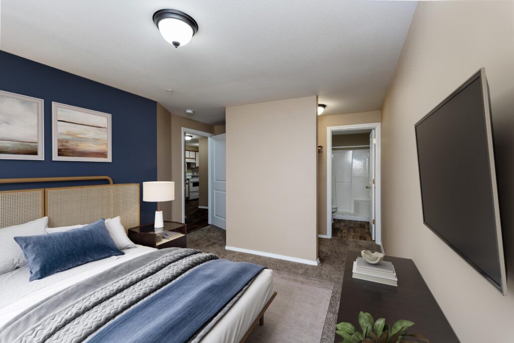 A modern bedroom with a bed, blue accent wall, two framed artworks, nightstand, lamp, large wall-mounted TV, and a door leading to a bathroom with a shower. A doorway shows a view into a kitchen area.