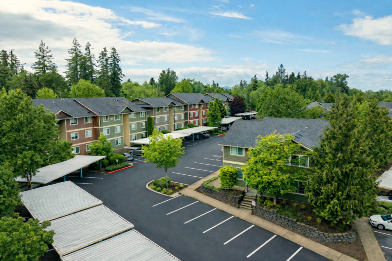 Aerial view of apartments in Salem OR, featuring multiple buildings, covered parking spaces, and surrounding trees under a partly cloudy sky. The parking lot appears mostly empty.