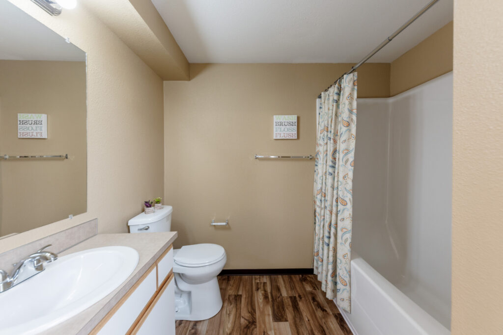 A clean bathroom with beige walls, a white sink and countertop, a toilet, and a bathtub with a patterned shower curtain. There is a mirror above the sink and a small sign on the wall above the toilet.