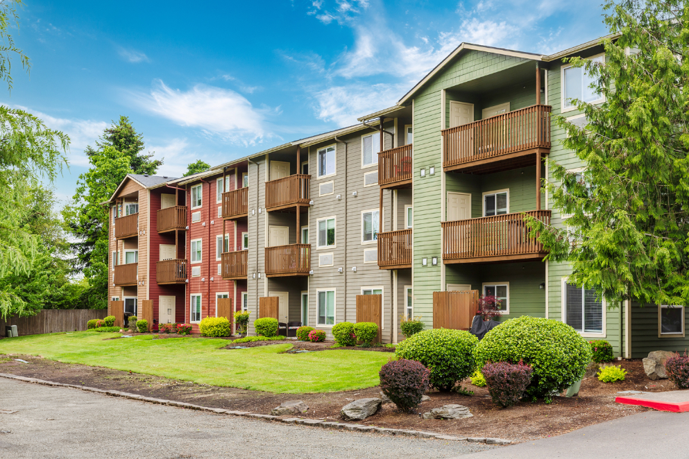 A modern apartment building with three stories, featuring wooden balconies and colorful siding in green, red, and brown—these apartments in Salem OR are surrounded by well-kept shrubs, lush grass, and large trees under a bright blue sky.