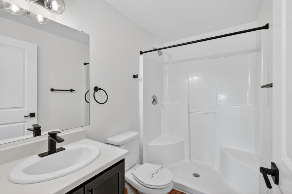 Modern bathroom with white walls, a sink with a black faucet, a toilet, and a walk-in shower with built-in seats and black fixtures. A large mirror hangs above the sink, and a towel ring is on the wall.