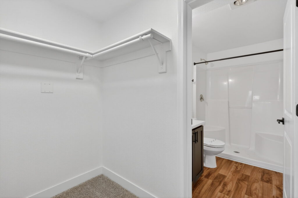 A walk-in closet with white walls and carpet leads to a bathroom featuring wood-look flooring, a white vanity, and a white shower-tub combination with a dark shower curtain rod.