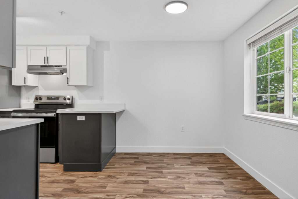 Modern kitchen with white cabinets, stainless steel oven, and dark lower cabinets. Hardwood flooring extends to an empty dining area by a large window with a view of green trees outside.