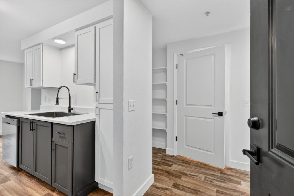 Modern kitchen with white upper cabinets, dark lower cabinets, a white countertop, a black faucet, and open shelving near a closed white door; wood-style flooring throughout the space.