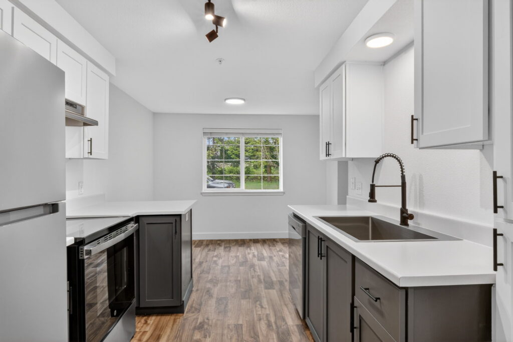 Modern kitchen with white cabinets, stainless steel appliances, dark lower cabinets, a large sink with a black faucet, white countertops, wood-style flooring, and a window letting in natural light.
