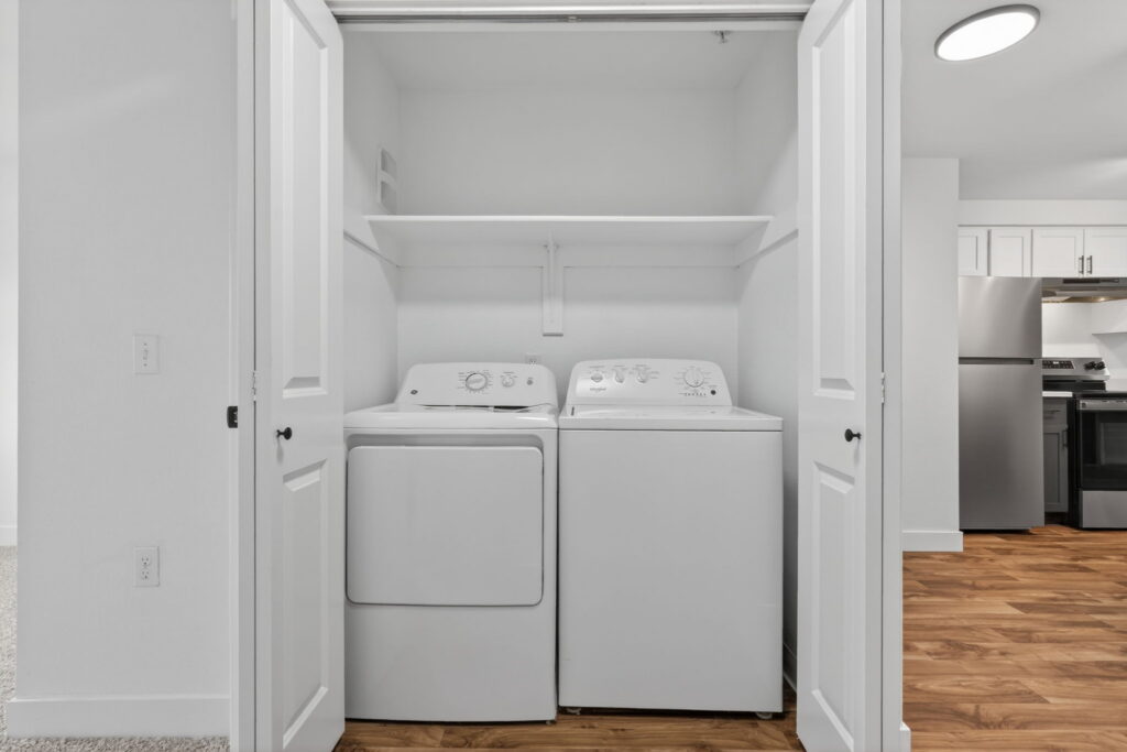 A laundry closet with white double doors open, revealing a white washing machine and dryer side by side beneath a white shelf, with a modern kitchen visible to the right.