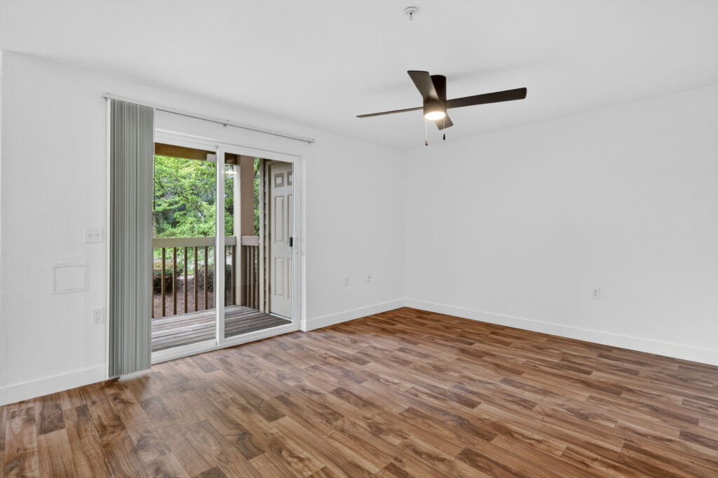 A bright, empty room with wood flooring, white walls, a ceiling fan, and a sliding glass door that opens to a covered balcony with a view of greenery outside.
