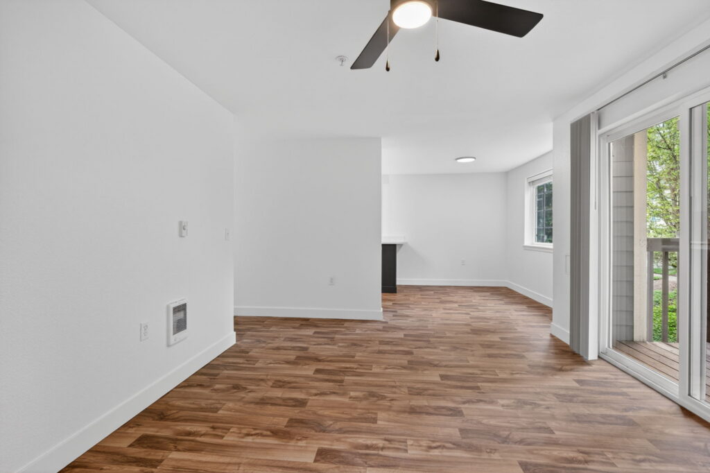 A bright, empty room with wood-look flooring, white walls, a ceiling fan, and a large sliding glass door leading to a small balcony with a view of green trees outside.