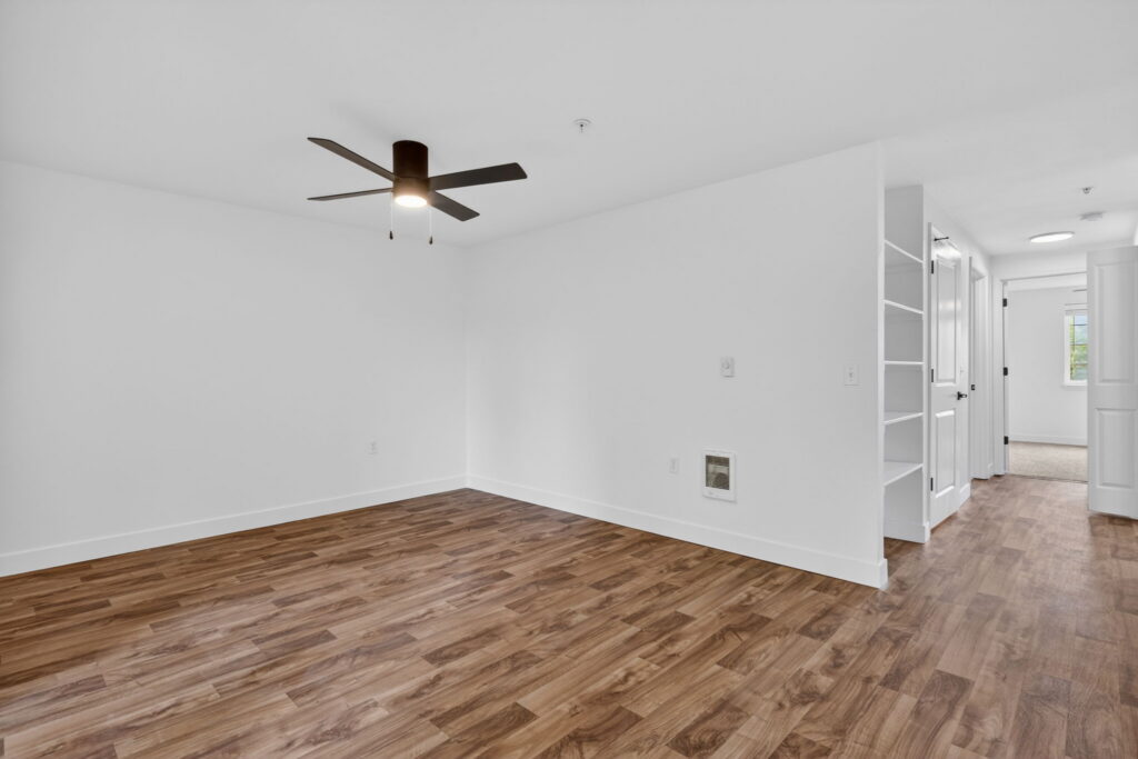 A bright, empty room with white walls, wood-style flooring, a ceiling fan with lights, open shelving, and a hallway leading to other rooms.
