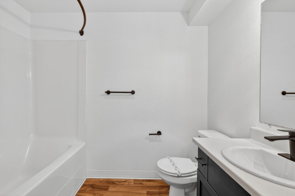A modern bathroom with a white bathtub and shower, white walls, a toilet with a plastic seal, dark fixtures, a white countertop with a sink, and wood-look flooring.