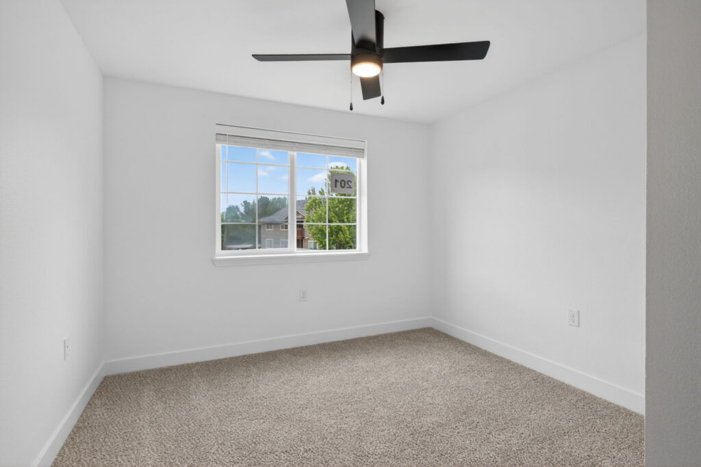 A bright, empty room with white walls, beige carpet, a large window letting in natural light, and a modern black ceiling fan with a light. Trees and a neighboring house are visible outside the window.
