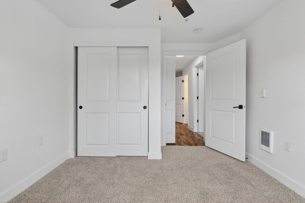 Empty bedroom with beige carpet, white walls, a ceiling fan, an open closet with sliding doors, and an open door leading to a hallway with wood flooring.