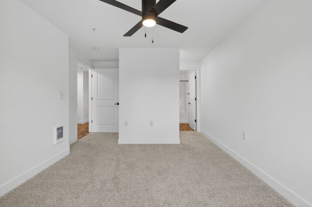 A bright, empty room with beige carpet, white walls, a black ceiling fan, and three white doors leading to other areas. The space appears clean and modern with minimalist design.
