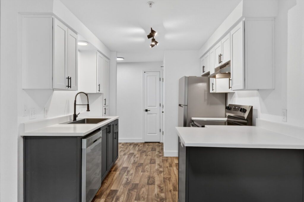 Modern kitchen with white upper cabinets, dark lower cabinets, stainless steel appliances, a farmhouse sink with a black faucet, under-cabinet lighting, and wood-look flooring. A white door is at the end of the room.