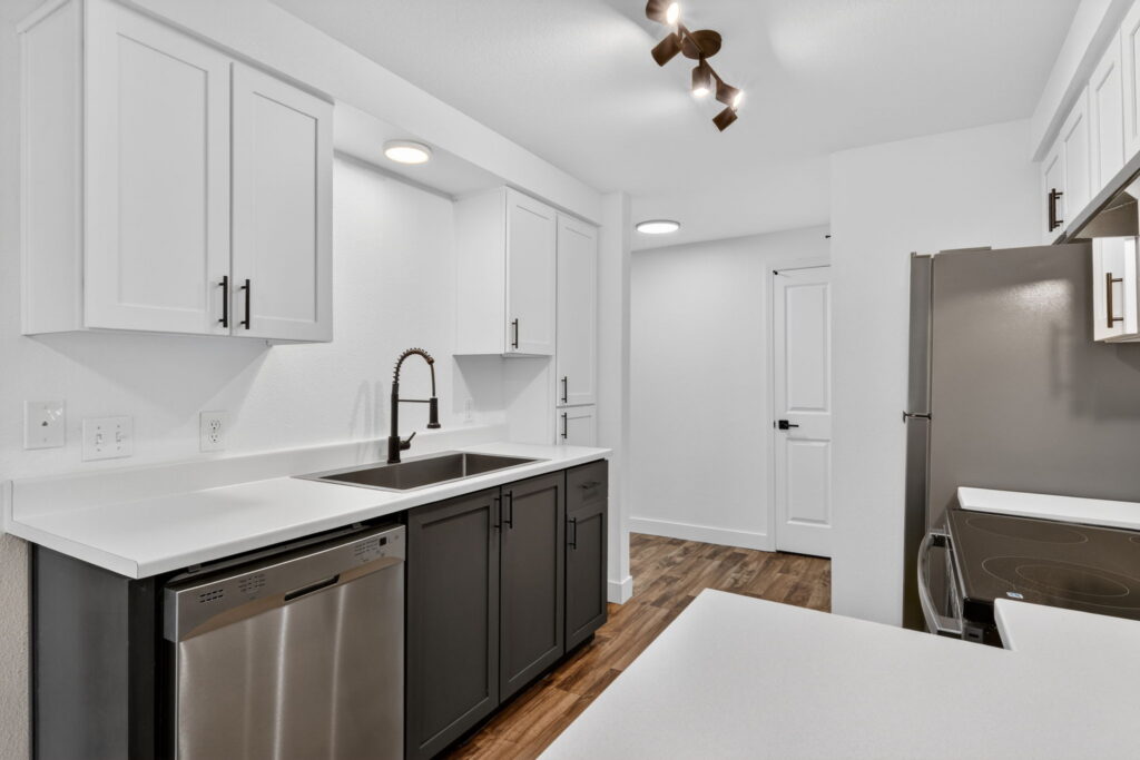 Modern kitchen with white upper cabinets, dark lower cabinets, stainless steel appliances, a double sink with a black faucet, white countertops, wood flooring, and minimal decor in a bright, clean space.