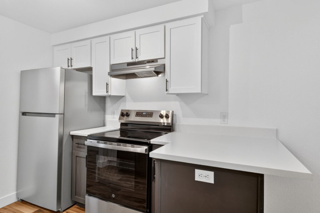 Modern kitchen with white cabinets, stainless steel refrigerator and oven, a range hood, and a white countertop with an L-shaped layout, featuring an electrical outlet on the counter’s side.