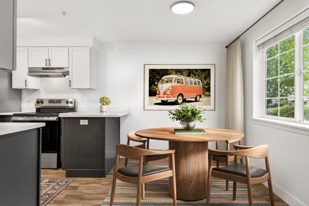 A modern kitchen and dining area with a round wooden table, four chairs, and a framed photo of a vintage van on the wall. Sunlight streams in through a window with beige curtains.