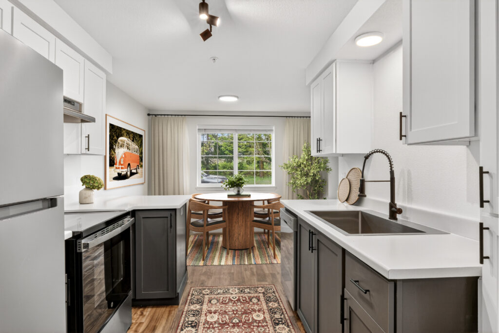 Modern kitchen with white cabinets, black appliances, a rug, and a dining area with a round table and chairs by a large window. A framed orange van photo hangs on the wall near green plants.